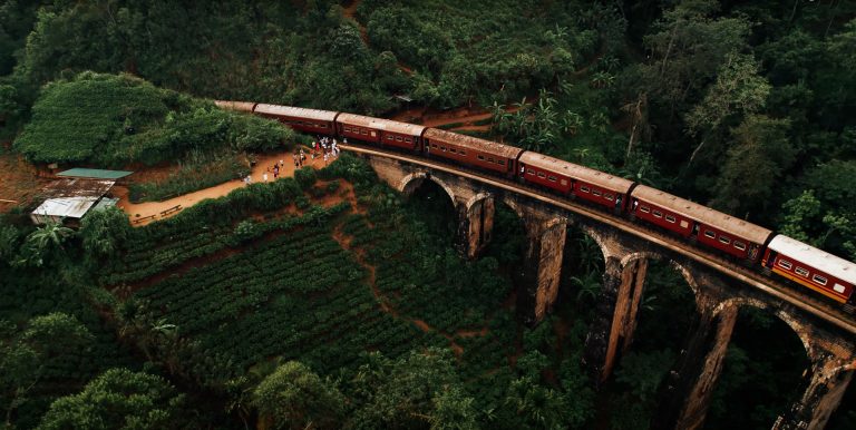 A red train crosses the iconic Nine Arches Bridge surrounded by lush green hills in Ella, Sri Lanka.