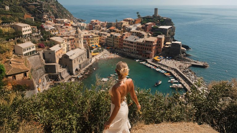 A woman overlooks the colorful seaside village of Vernazza in Cinque Terre, with its harbor and pastel houses by the Ligurian Sea.
