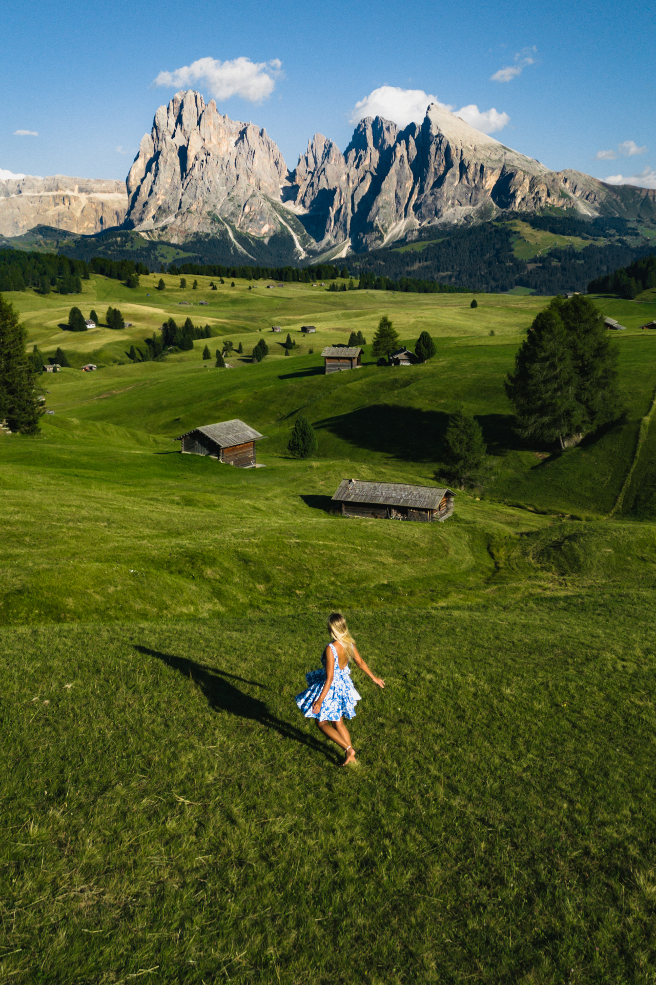 Woman in blue dress walking across green meadows of Alpe di Siusi with Dolomites peaks behind.