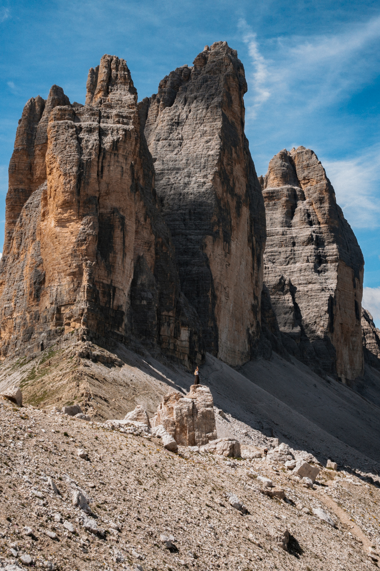 Person standing on rocks at the three peaks beneath towering Dolomites cliffs with bright blue sky above.