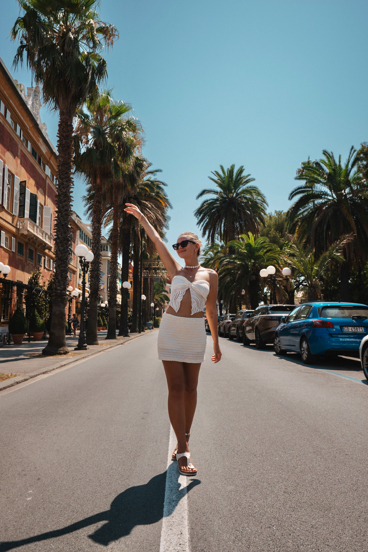 Woman walking down a palm-lined street in Sestri Levante on a sunny day, wearing a white outfit.