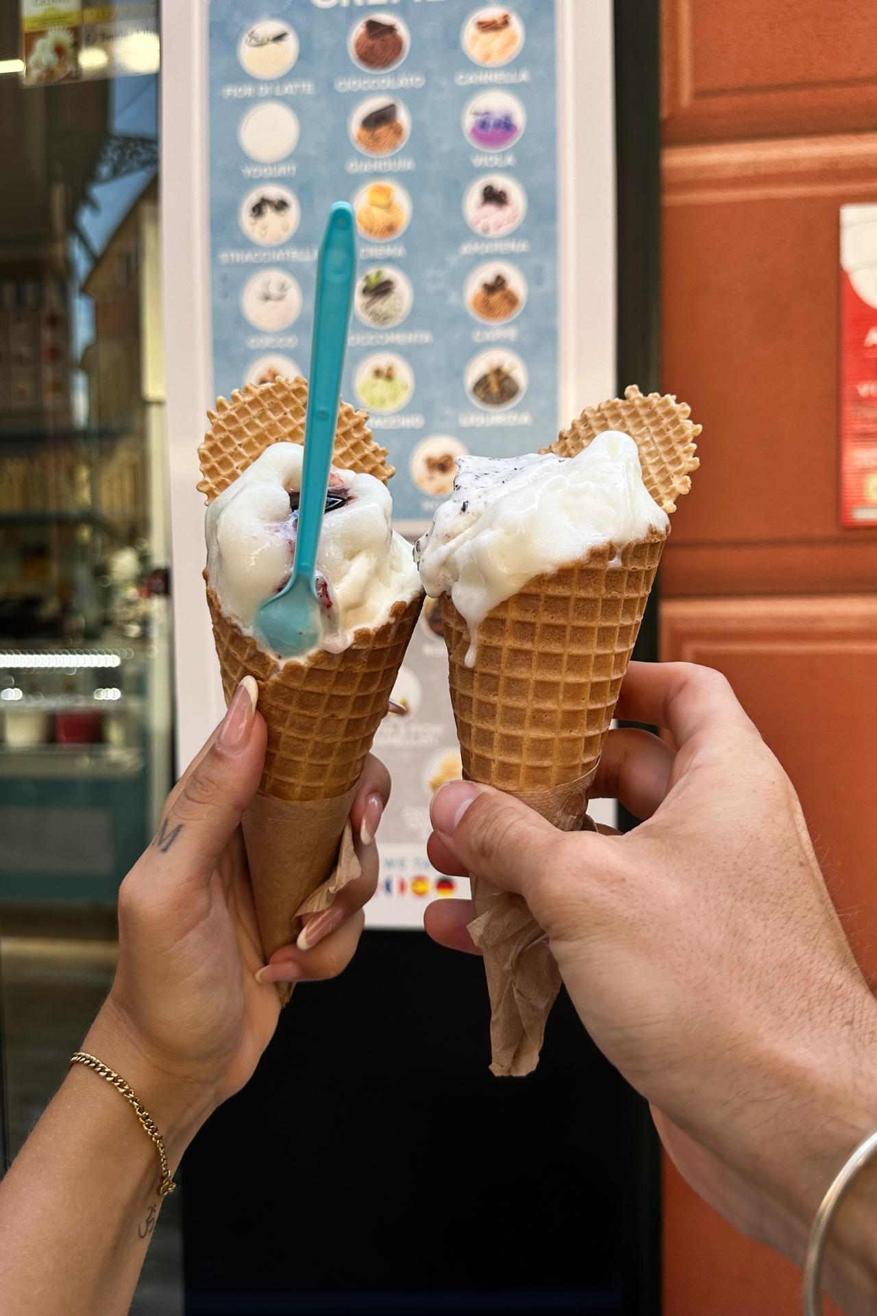 Two hands holding gelato cones in Sestri Levante old town with a flavor menu in the background.