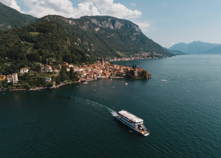 Aerial drone view of Varenna town and Lake Como shoreline with colorful houses, marina, and surrounding mountains.