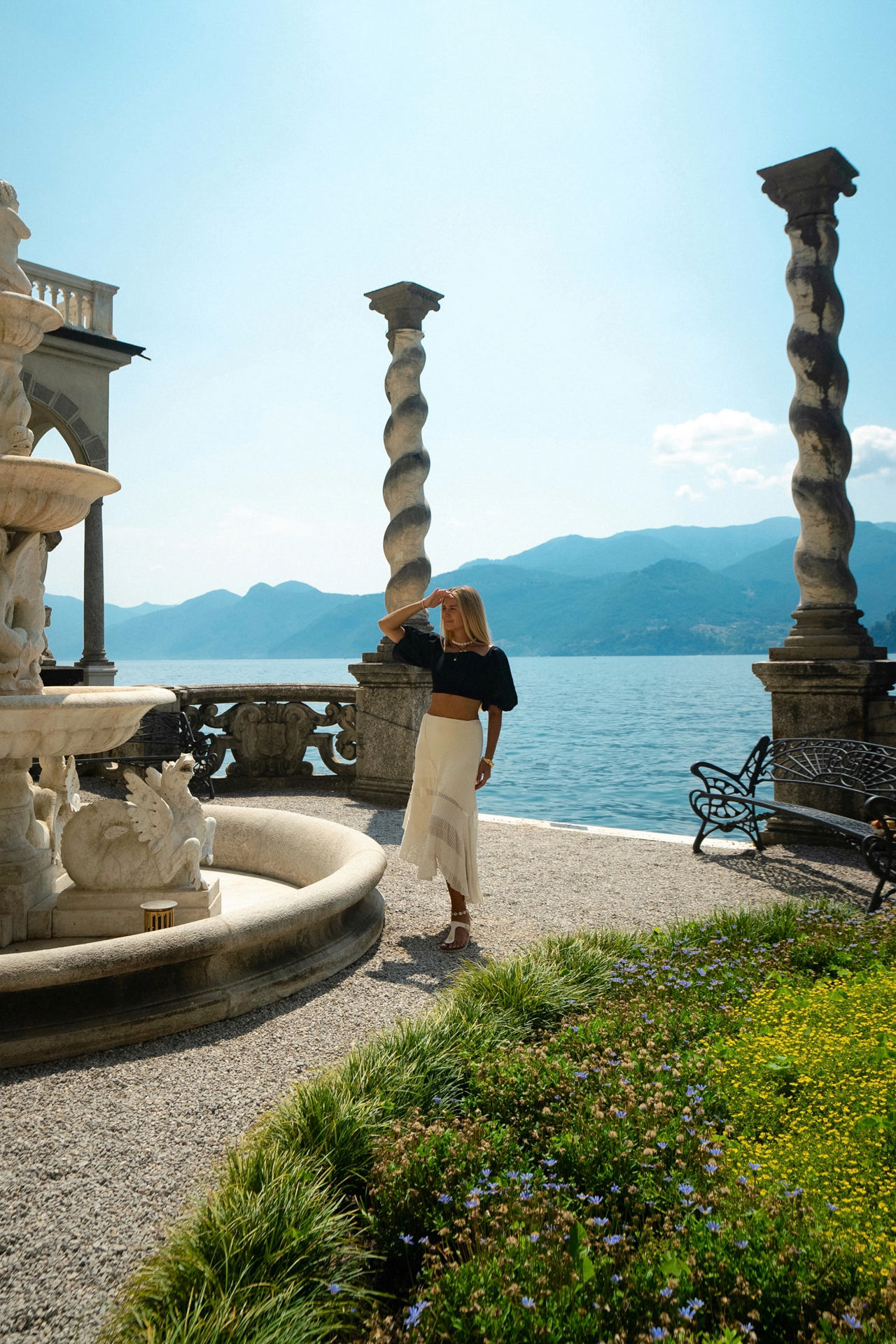 Woman standing near a fountain with carved statues and twisted columns overlooking Lake Como at Villa Monastero, Varenna, Italy.