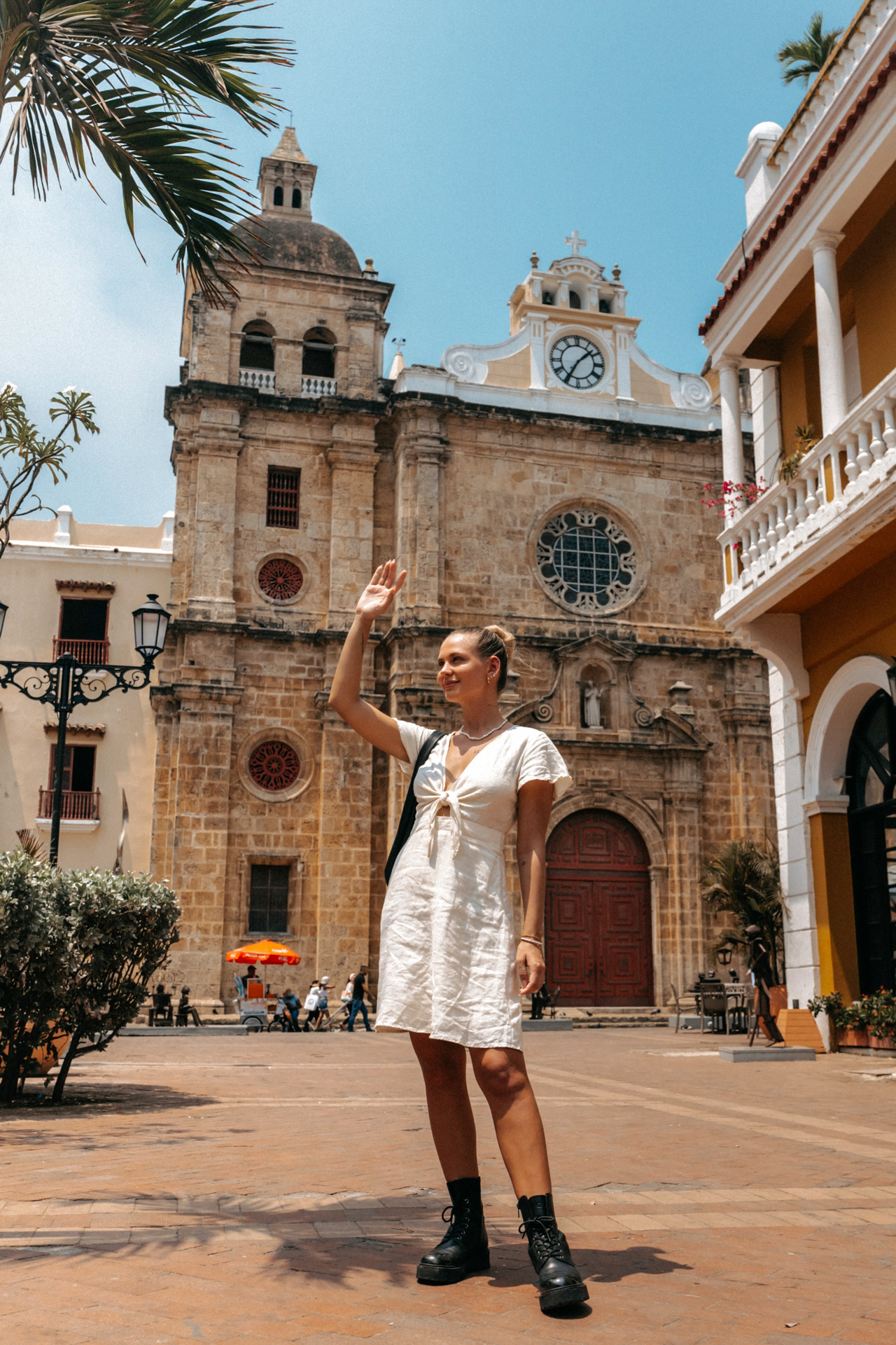 Woman in a white dress raising her hand in front of the historic Cartagena Cathedral in Colombia.
