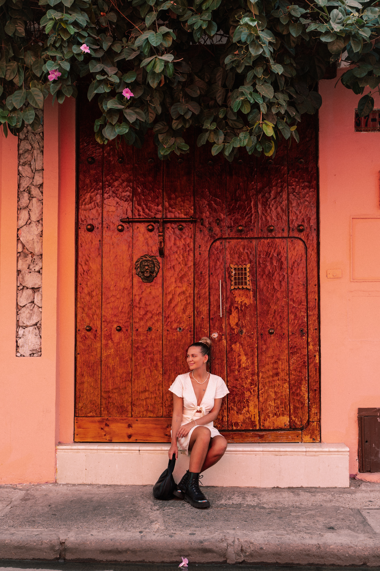Woman sitting in front of a large wooden colonial door framed by pink walls and flowers in Cartagena, Colombia.