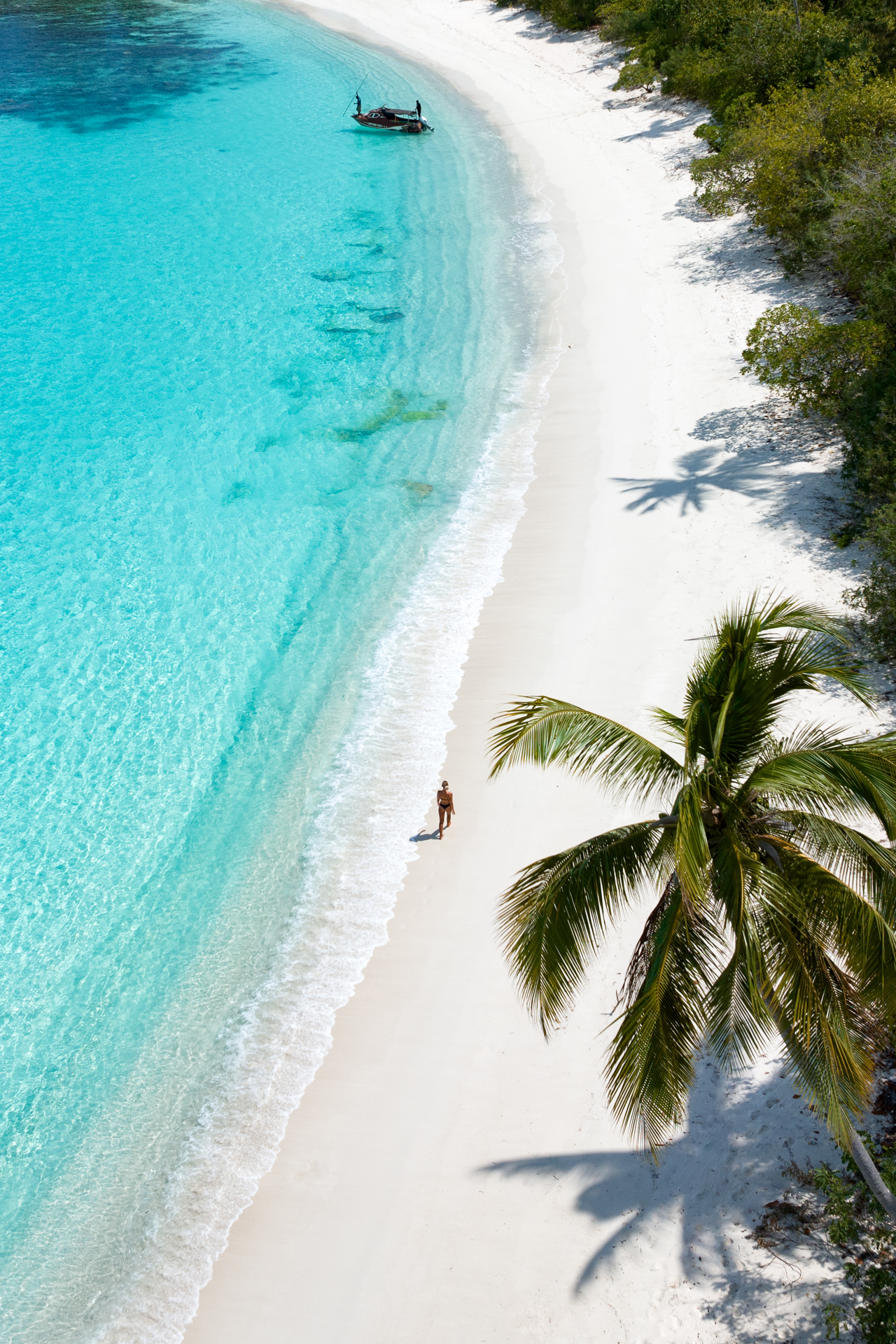 A woman walks along a white sandy beach with turquoise waters and a palm tree casting shadows.