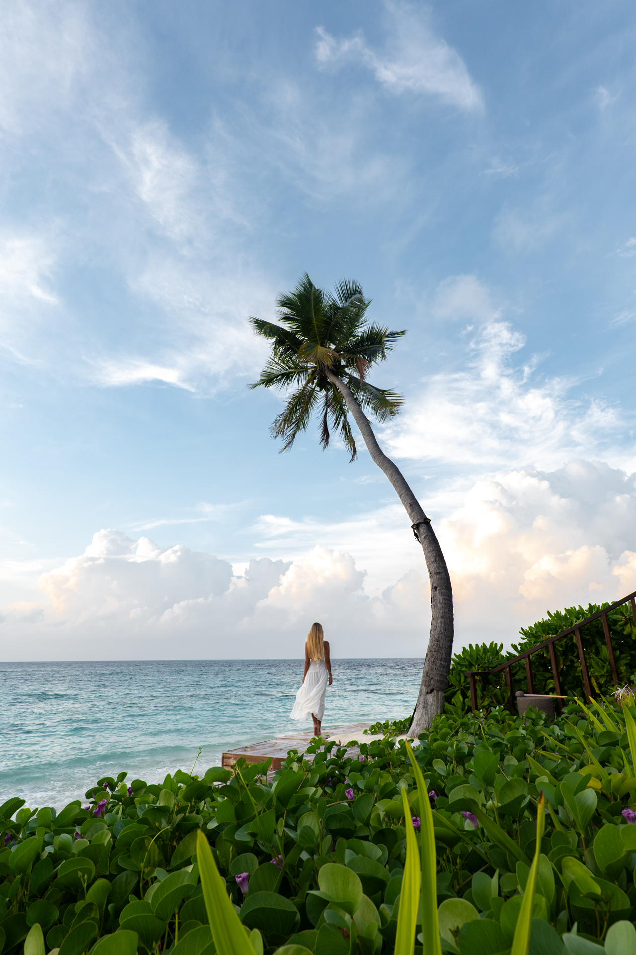 Woman in a white dress walking by the ocean under a leaning palm tree in the Maldives, with blue skies and lush greenery.