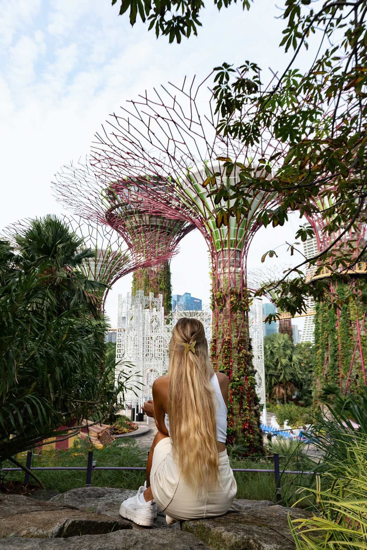 Woman sitting on a rock, facing the Supertree Grove at Gardens by the Bay in Singapore, surrounded by lush greenery.