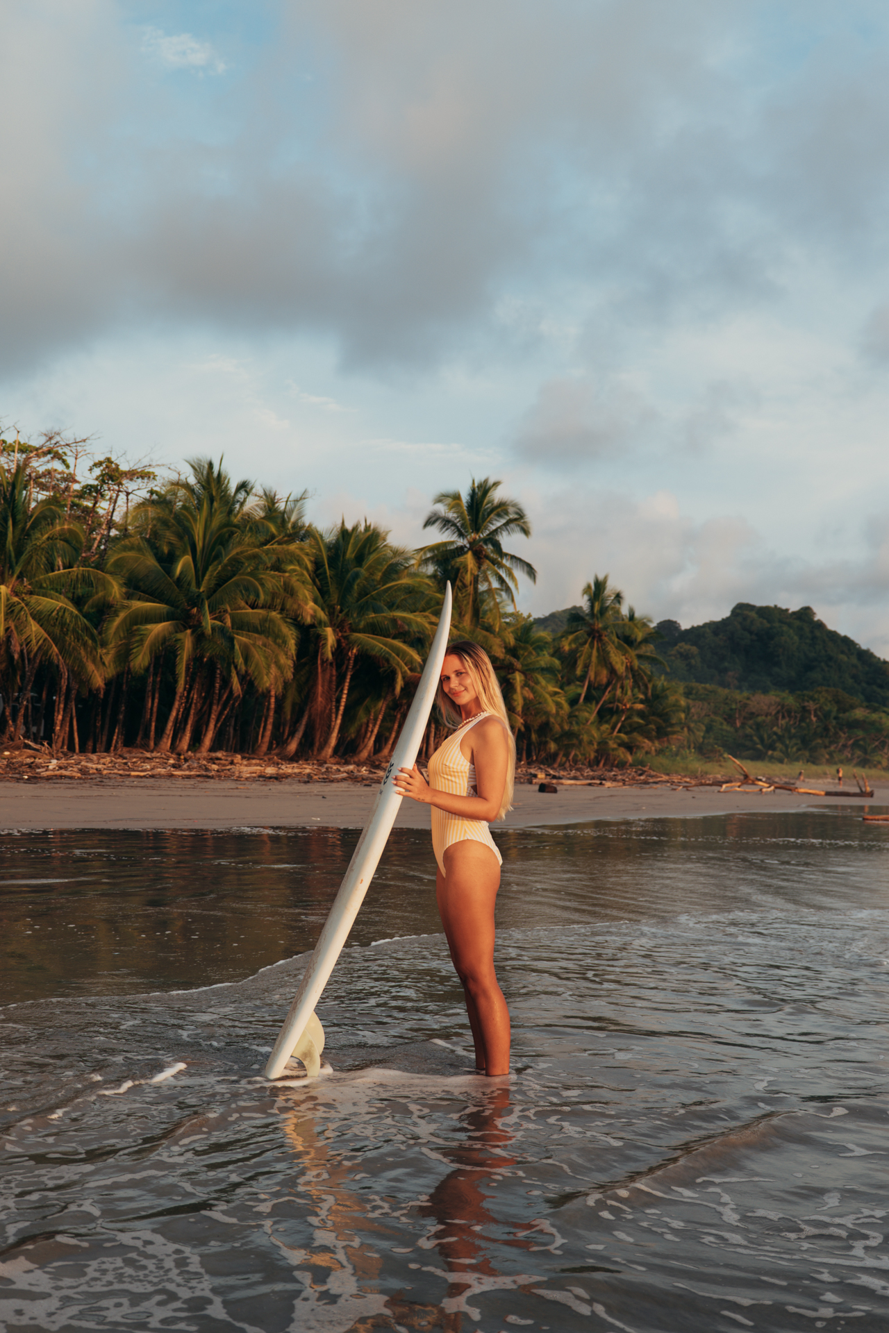 Woman holding a surfboard while standing in shallow ocean water at sunset in Santa Teresa, Costa Rica