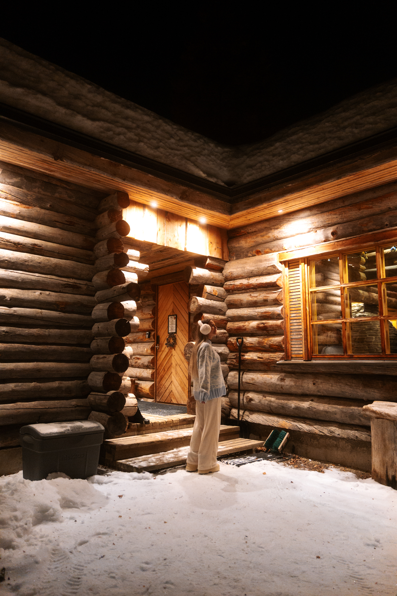 Pyhä ALT text: Woman standing outside a traditional wooden cabin at night in Pyhä, Finland surrounded by snow