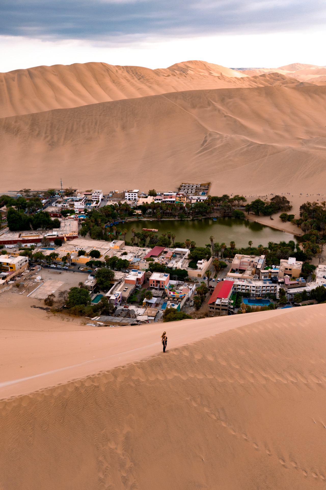 Woman walking on sand dunes overlooking Huacachina oasis and village, Peru