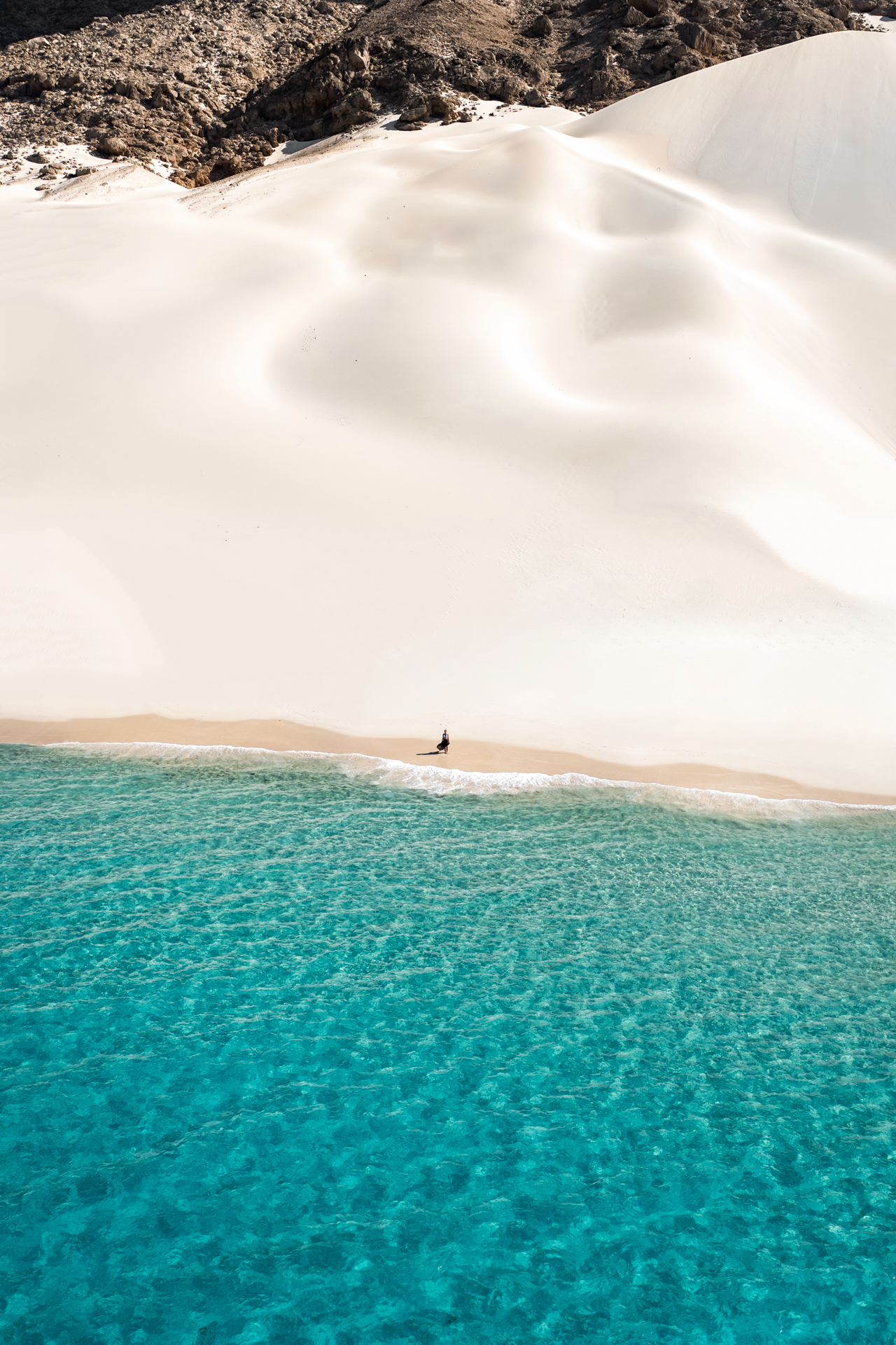 Person walking along turquoise shore beneath towering white dunes at Arher Sand Dunes.