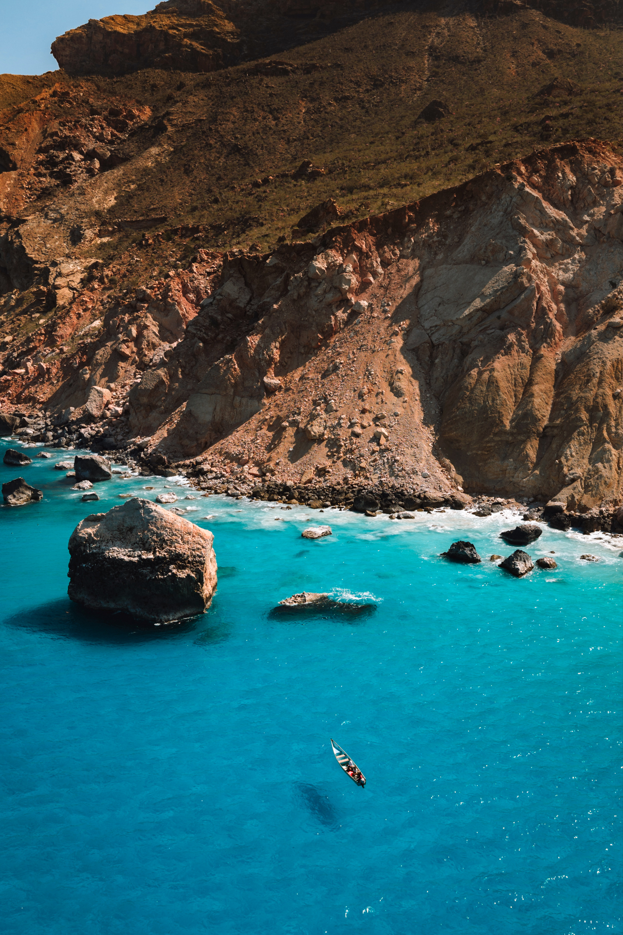 A small boat floats on the crystal-clear turquoise waters beneath rugged coastal cliffs on Socotra Island.