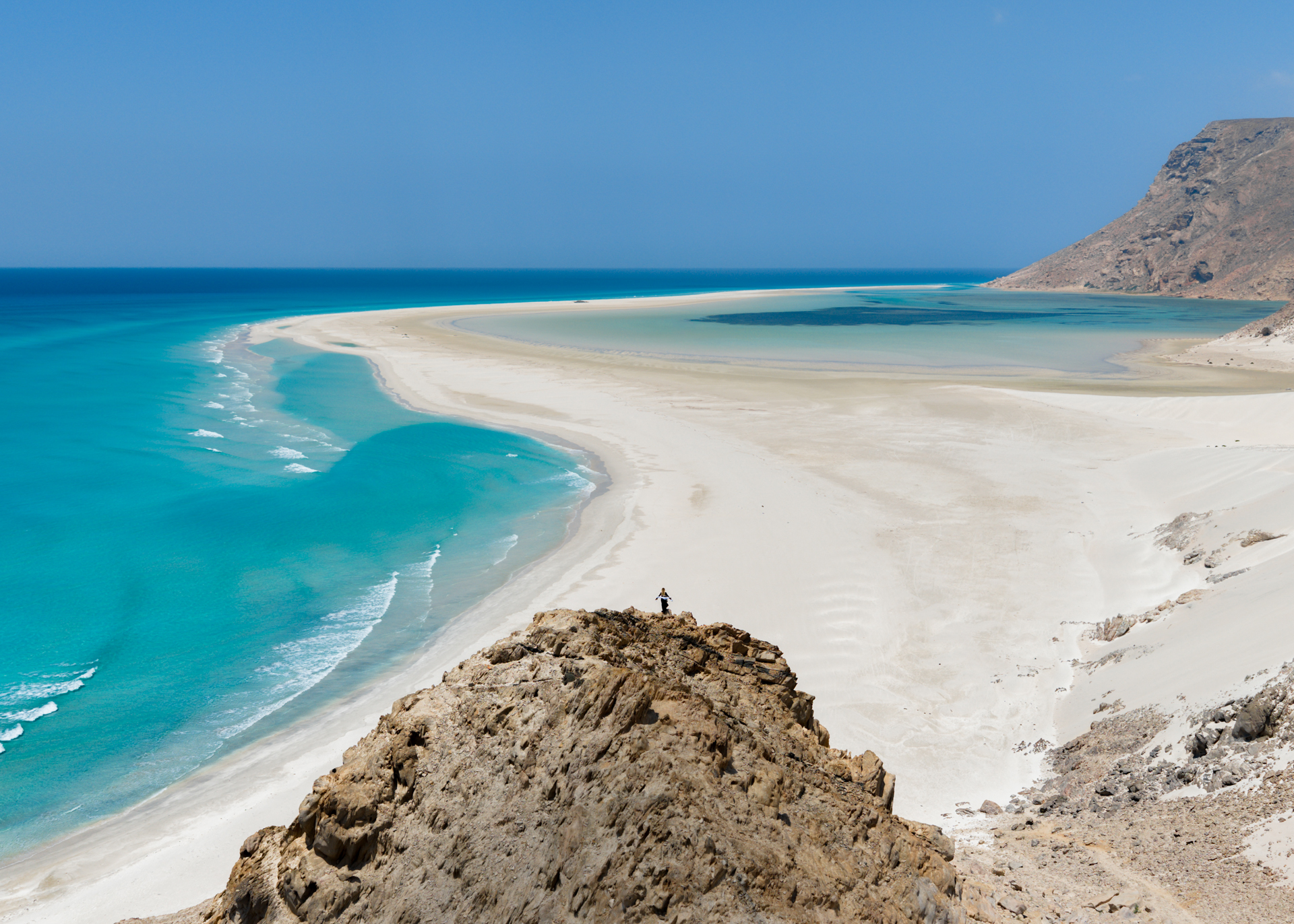 A person standing on a rocky cliff overlooking the stunning turquoise waters and curved sandbar of Detwah Lagoon on Socotra Island.