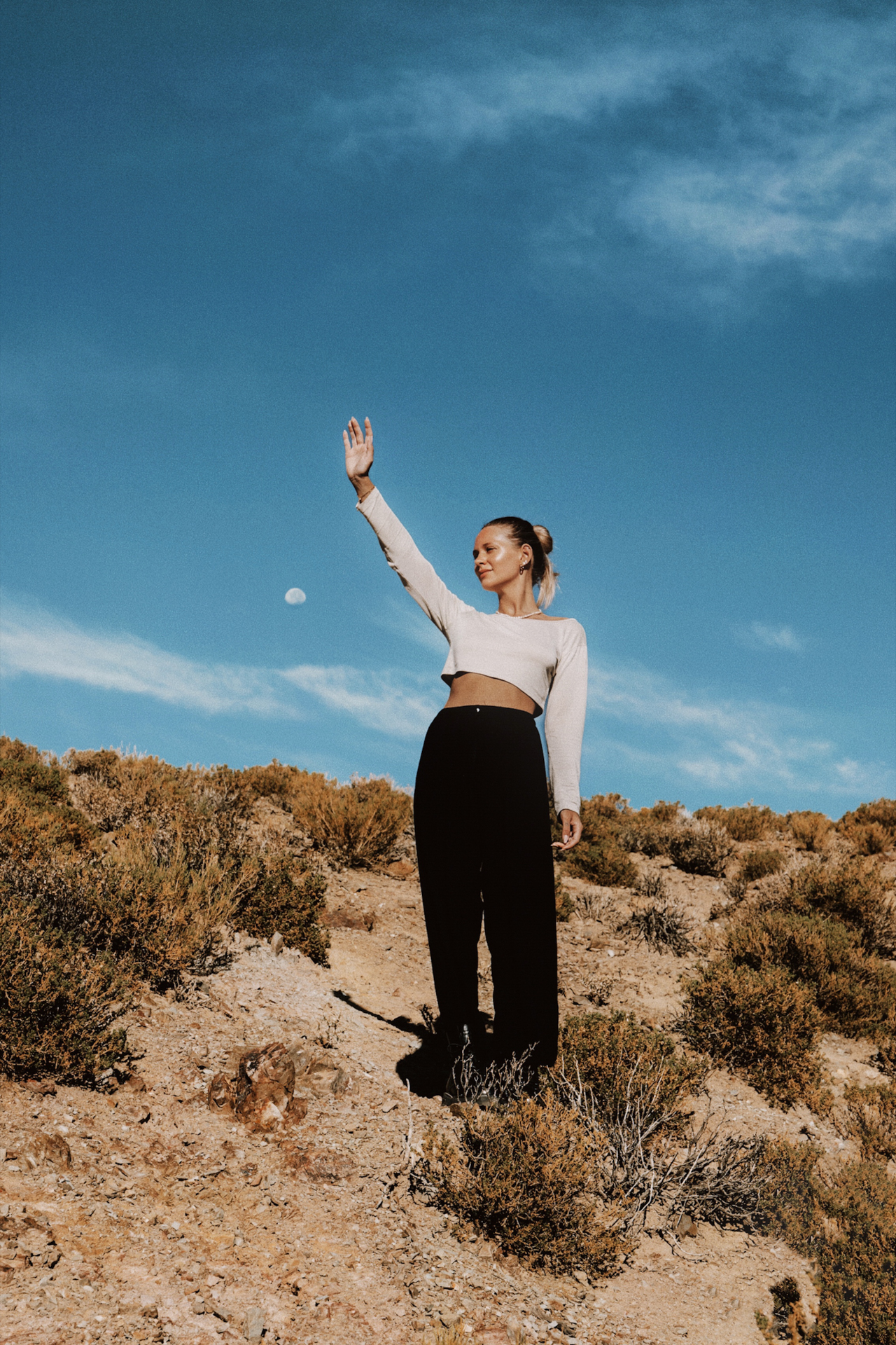 Woman standing on a dry hillside under a clear blue sky, raising her hand playfully toward the moon in Bolivia.