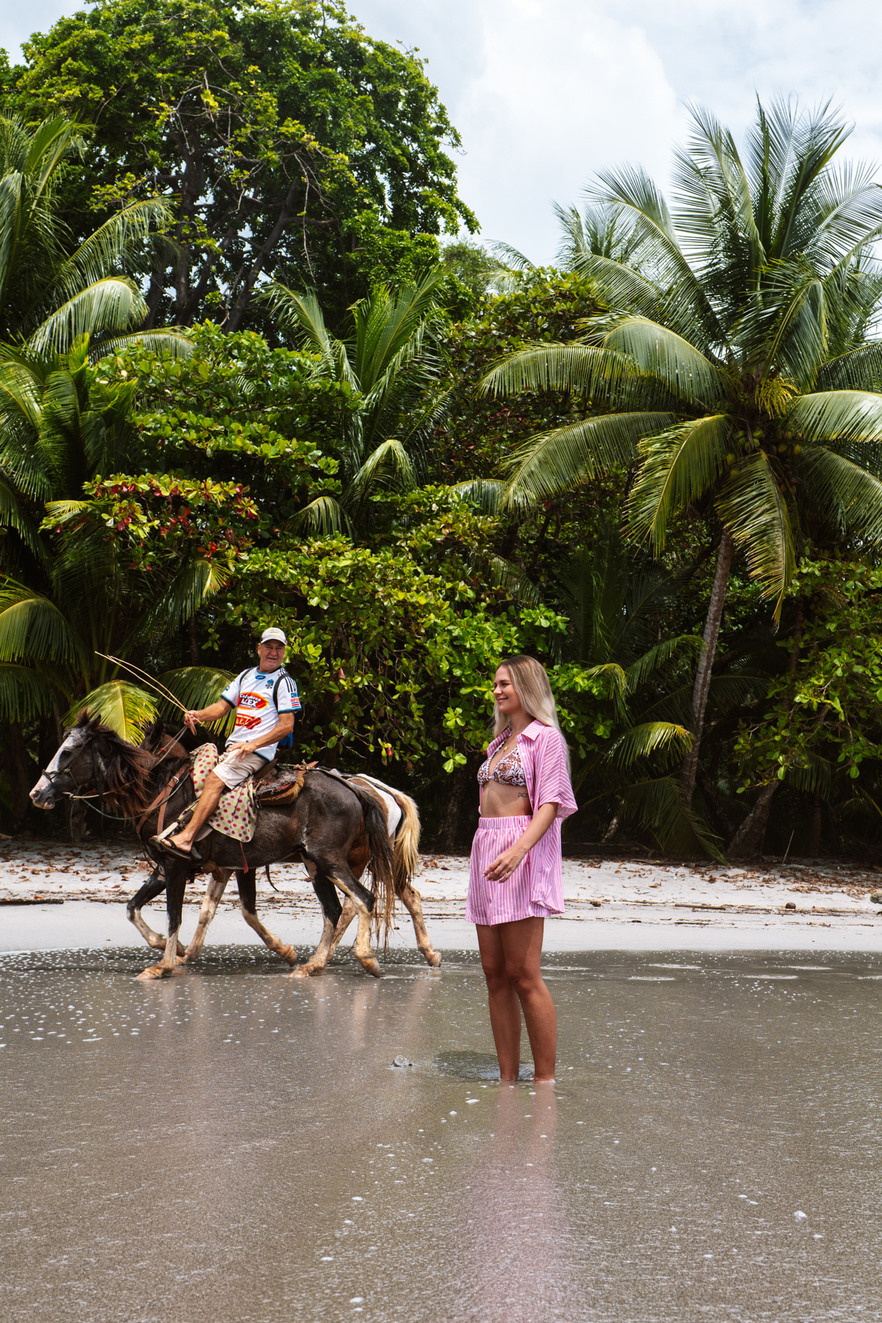 Woman standing in the ocean on tropical beach in Costa Rica as man rides horse along shore