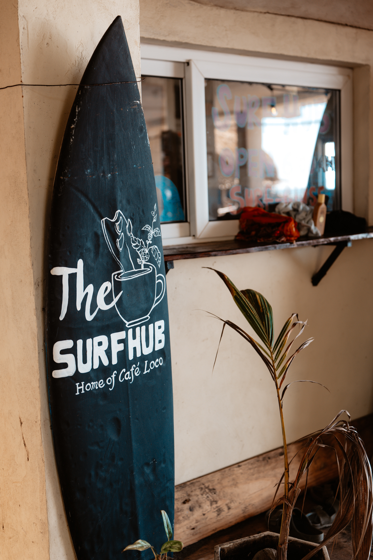 Surfboard sign outside The Surf Hub café in El Zonte with small plant and window counter
