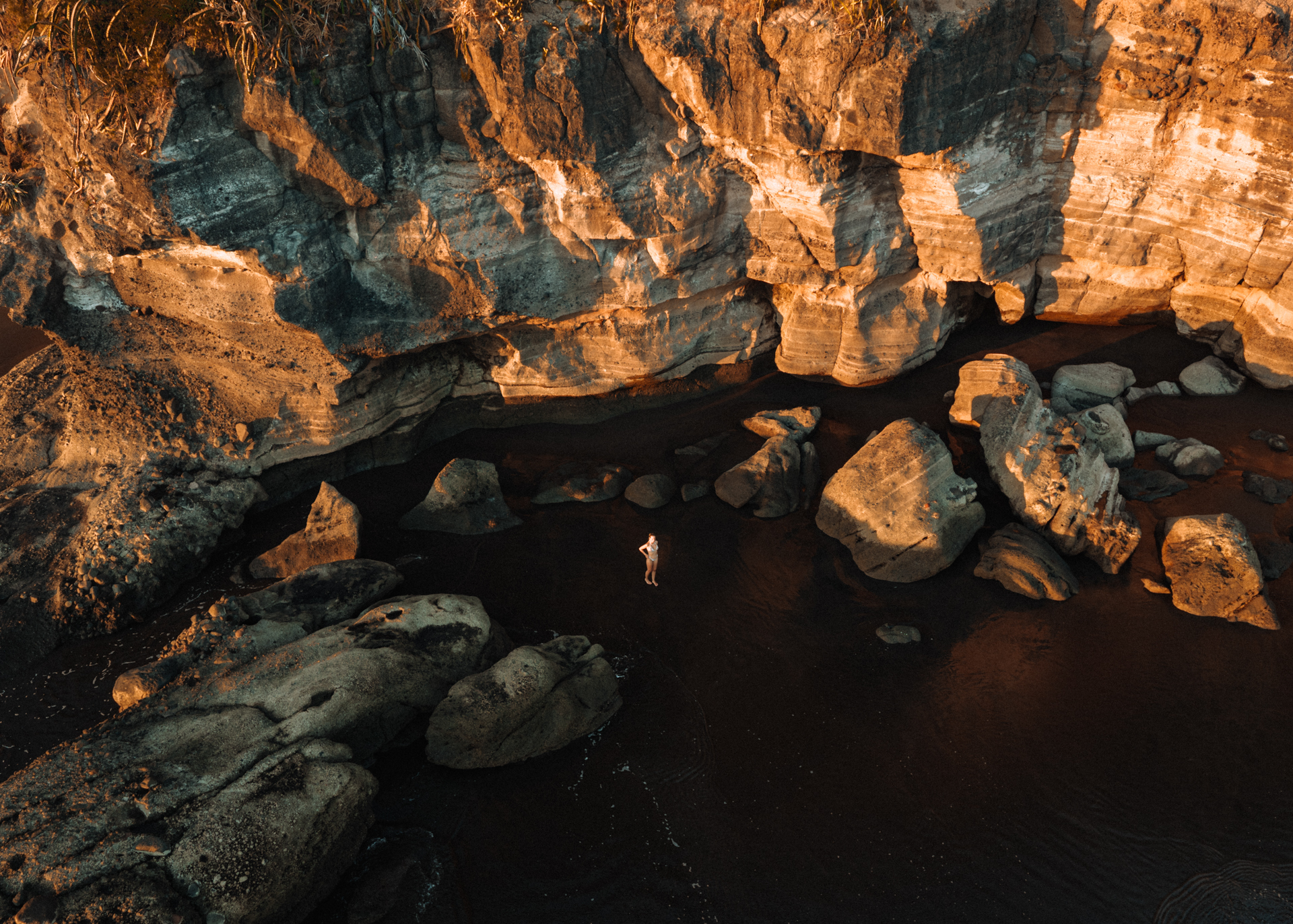 Aerial view of a person standing in a dark river surrounded by golden cliffs in El Zonte, El Salvador at sunset