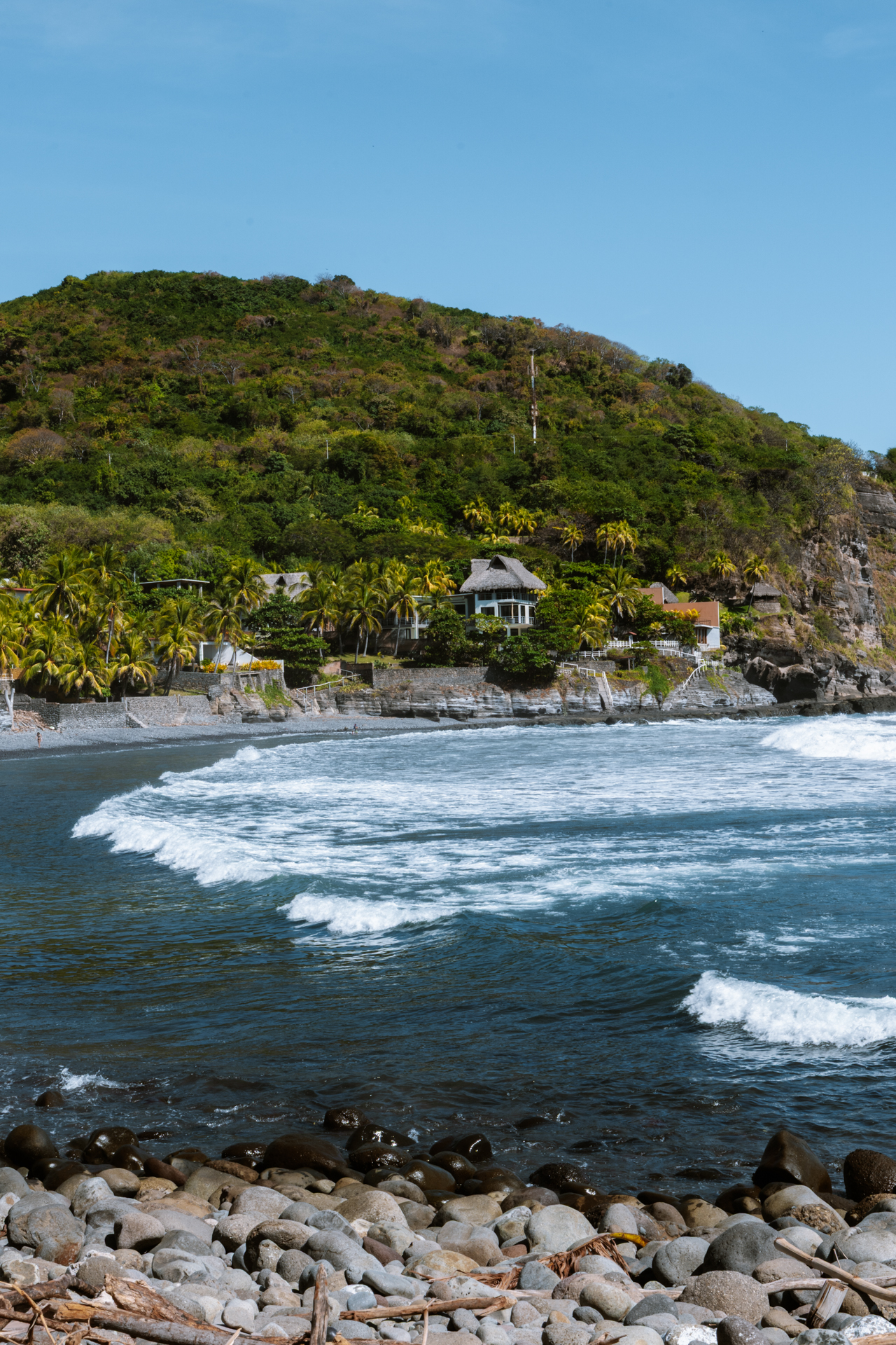 Rocky beach and ocean waves at El Zonte with tropical hillside houses and palm trees in El Salvador.