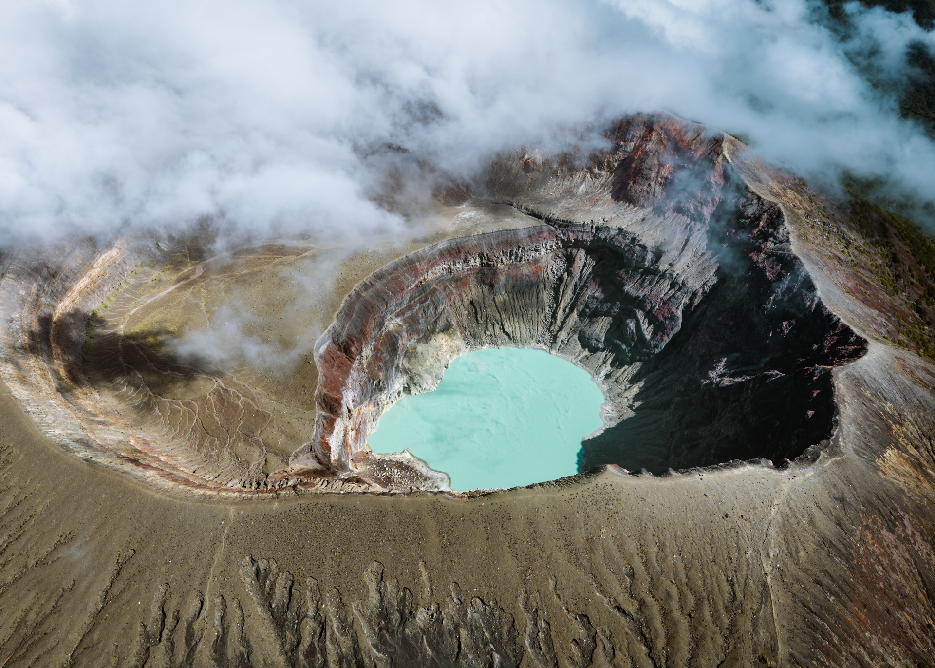 Aerial view of a volcanic crater with a turquoise lake surrounded by rugged terrain and clouds