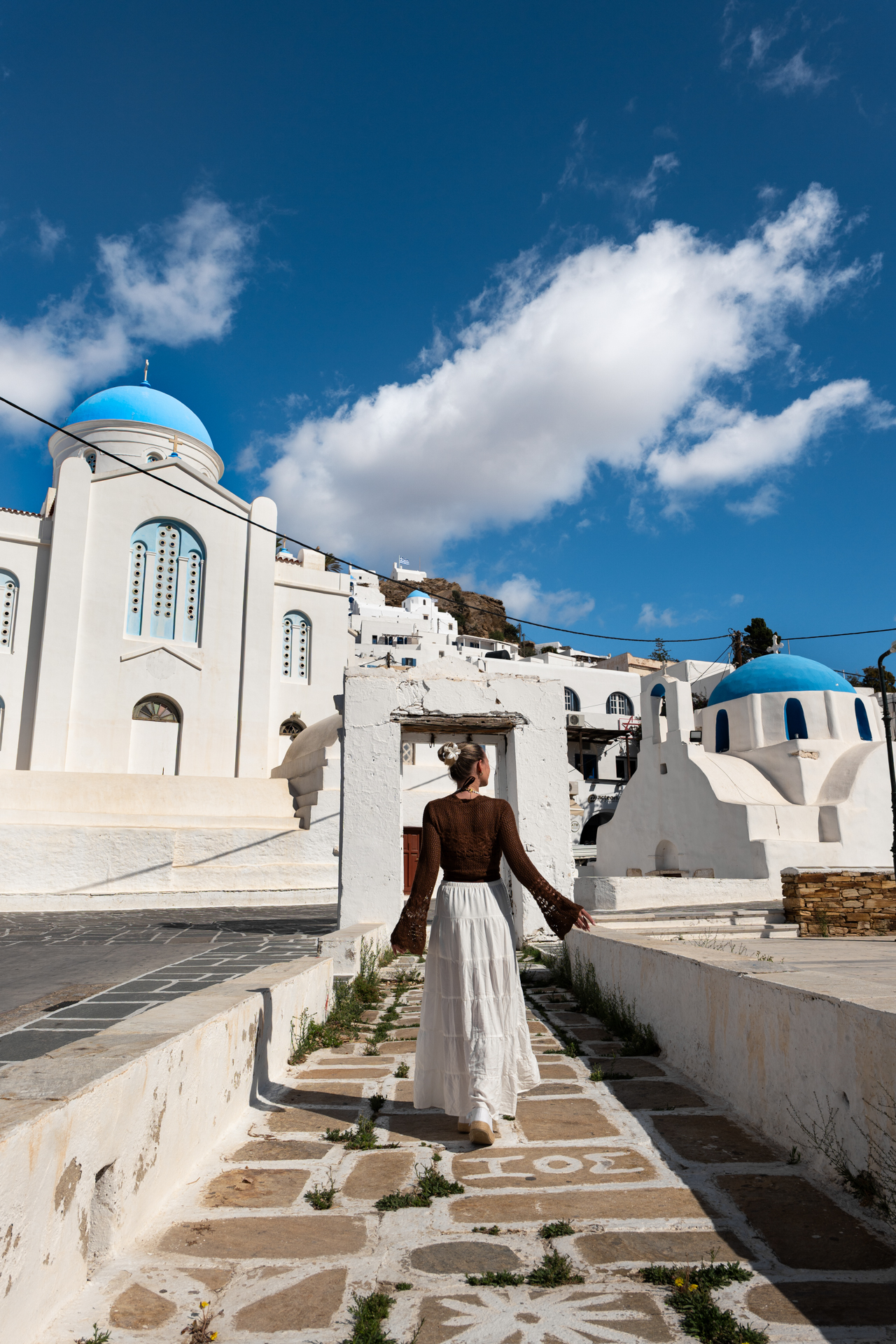 Woman walking through white alley with blue-domed church in Chora, Ios