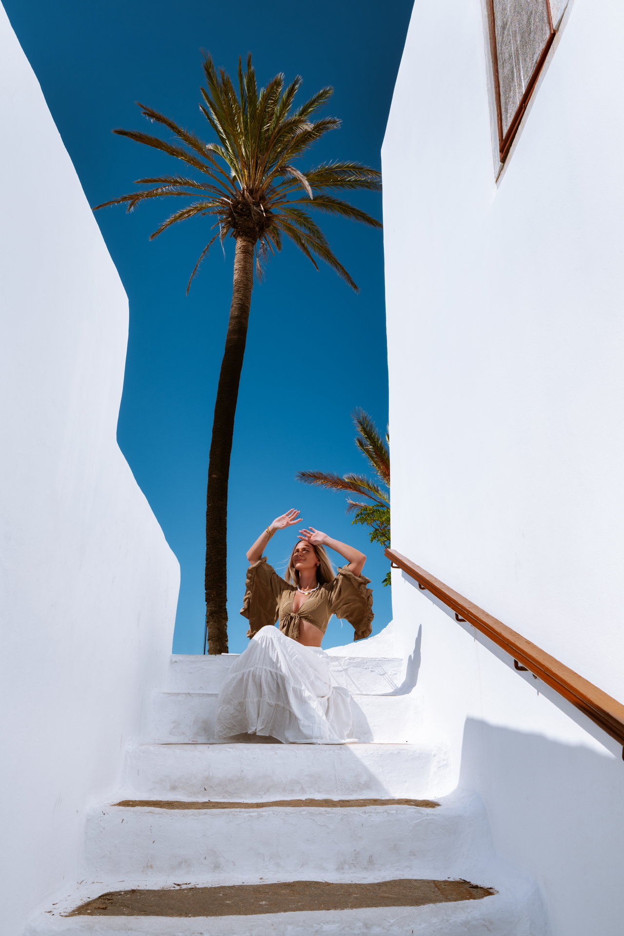 Woman sitting on white stairs in Chora, Ios with palm tree and blue sky