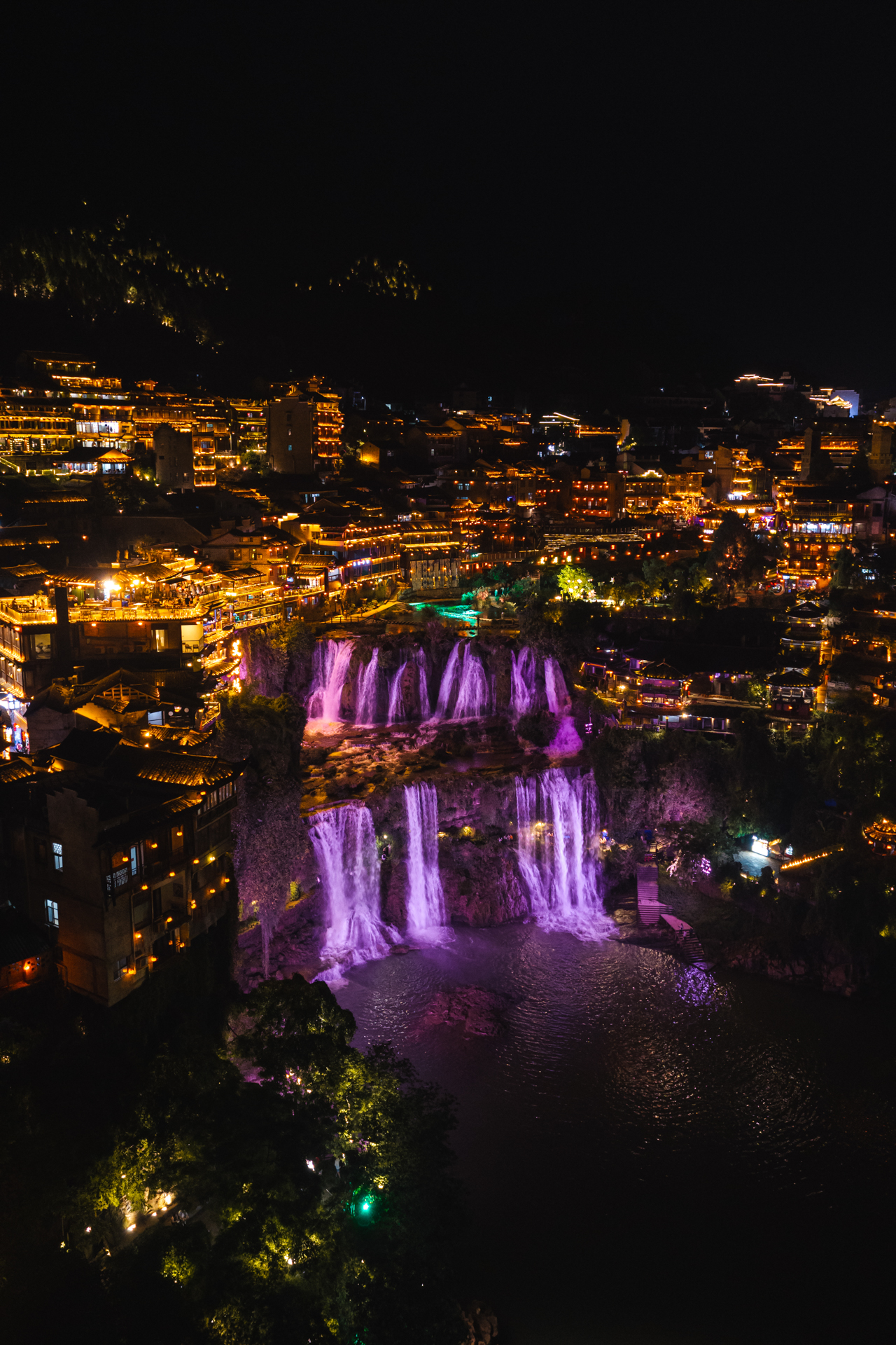 Furong Ancient Town Furong Ancient Town in Hunan, China, glowing at night with illuminated waterfalls, traditional buildings, and colorful riverside lights.