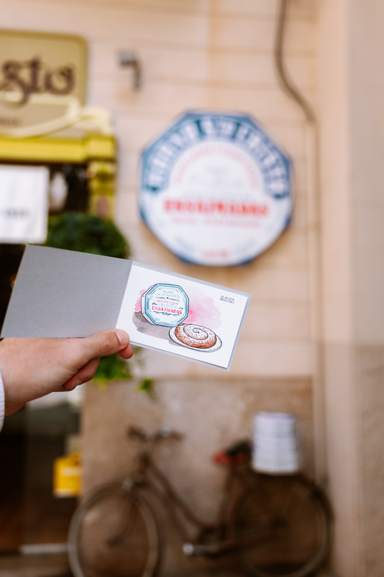 Hand holding an illustrated ensaïmada card in front of a traditional bakery in Palma de Mallorca, Spain.