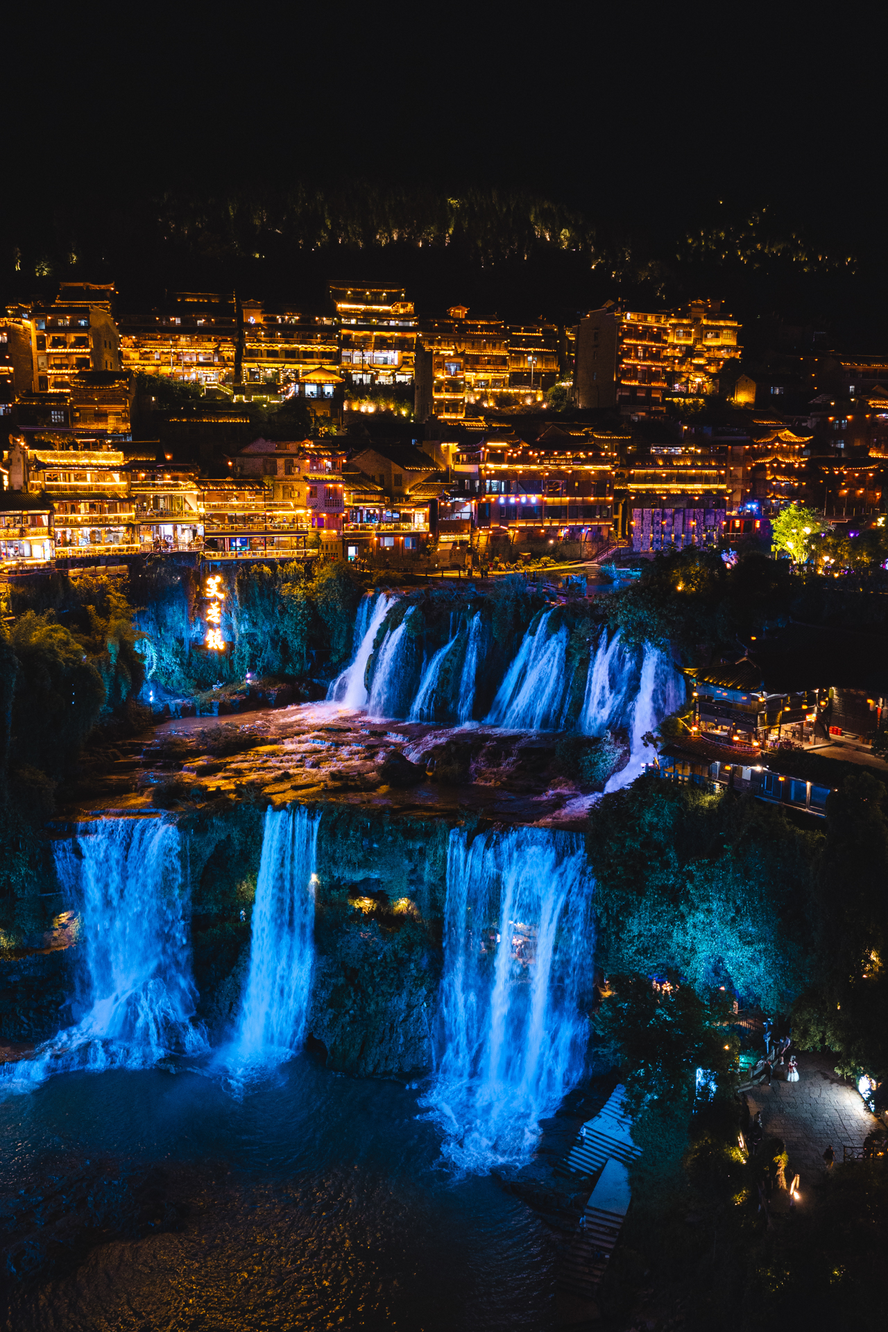 Furong Ancient Town glowing at night with blue-lit waterfalls and traditional buildings.