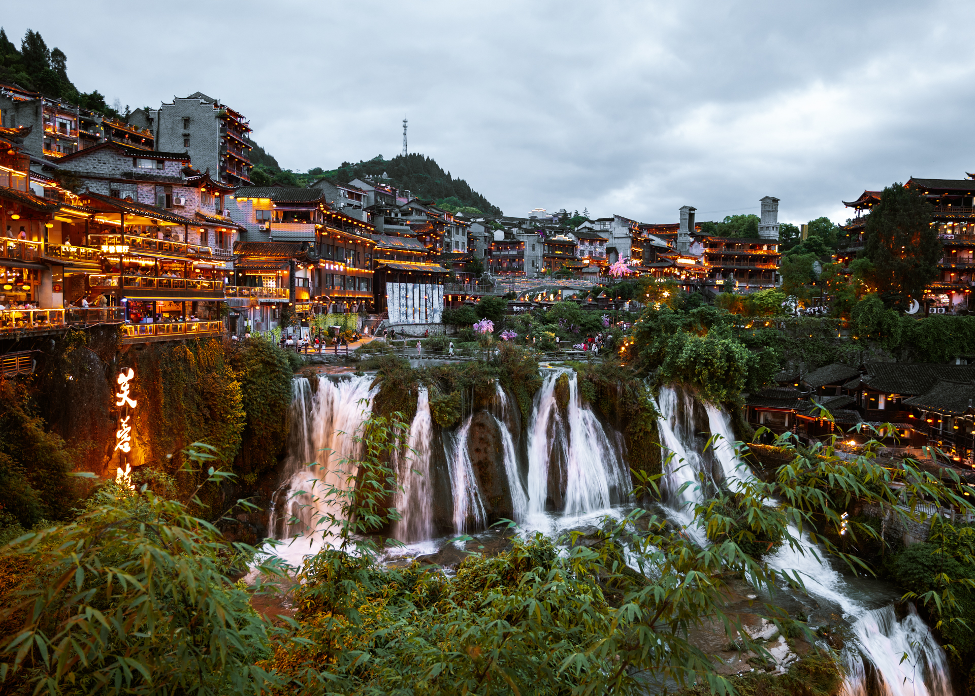 Furong Ancient Town waterfall surrounded by illuminated traditional buildings at dusk.