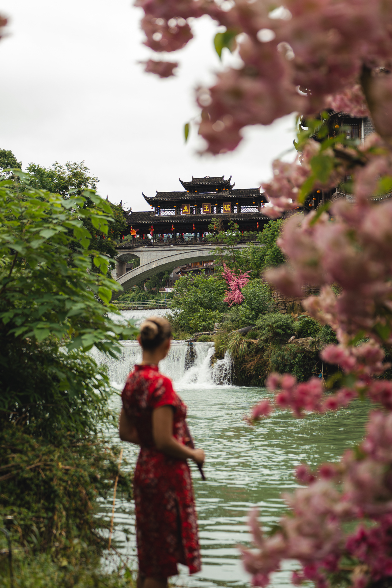 Woman in a red dress overlooking Furong Ancient Town bridge and waterfall in Hunan, China.
