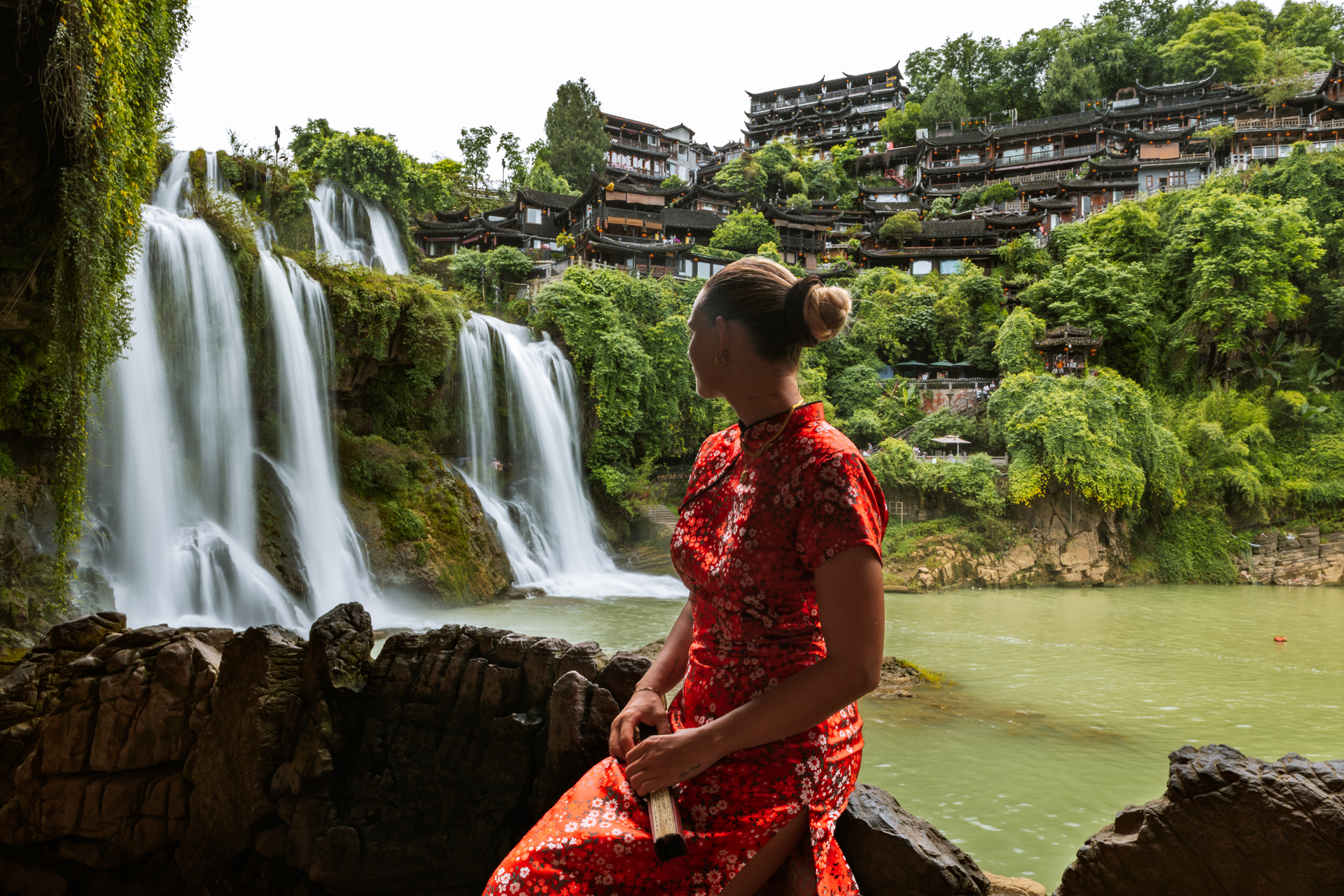 Woman in a red dress overlooking the waterfall and traditional buildings of Furong Ancient Town.