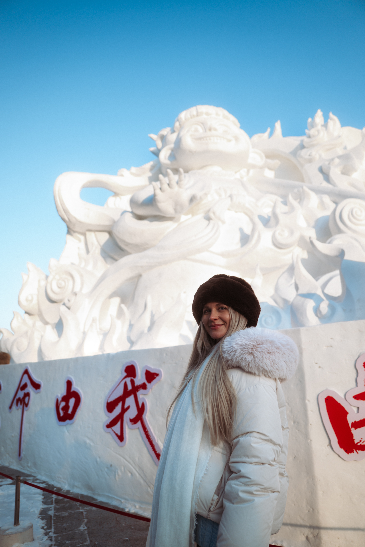 Woman posing in winter clothing beside a large snow sculpture in Harbin, China.