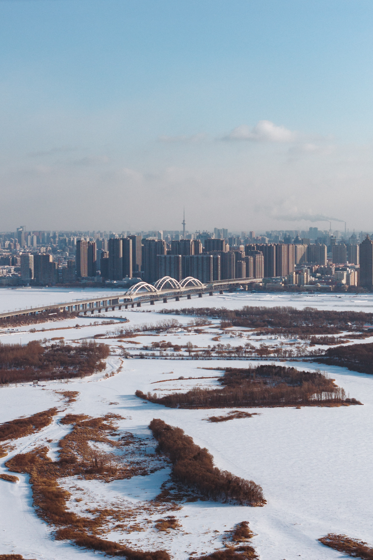 Frozen Songhua River and Harbin skyline with a bridge crossing the snowy winter landscape.