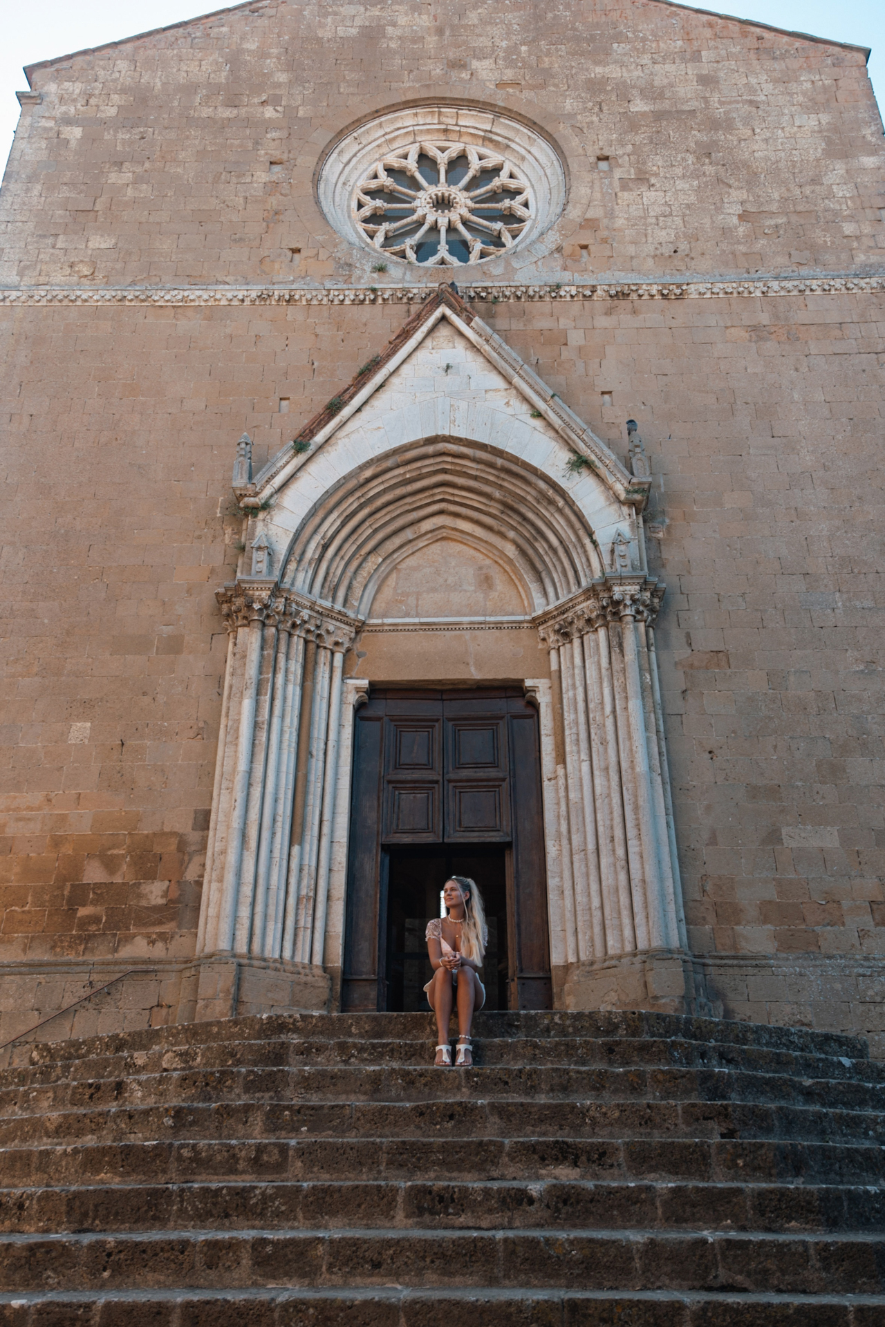 Woman sitting on the steps of a historic church with a large arched doorway in Monticchiello, Tuscany.